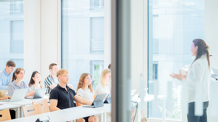 Man blickt seitlich in einen hellen Vorlesungsraum. Eine junge Professorin erklärt einigen Studierenden einen Sachverhalt. Im Hintergrund sieht man große Fenster, durch die ein weiteres Gebäude zu sehen ist.