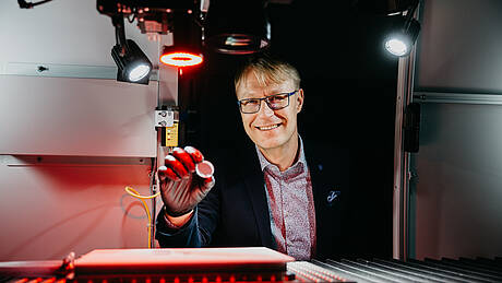 Prof. Dr. Thomas Schwarzbäck, der im Bereich Lasermaterialbearbeitung forscht und lehrt, sitzt an einem Versuchsaufbau, der rot angeleuchtet wird.