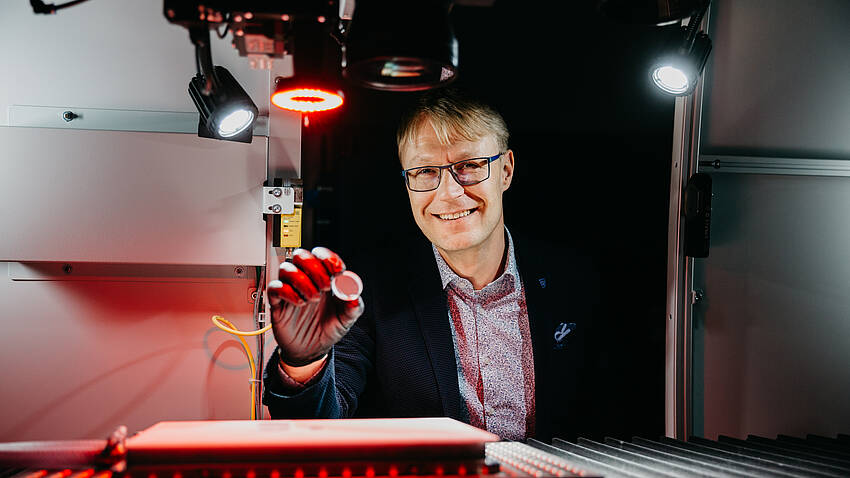 Prof. Dr. Thomas Schwarzbäck, der im Bereich Lasermaterialbearbeitung forscht und lehrt, sitzt an einem Versuchsaufbau, der rot angeleuchtet wird.