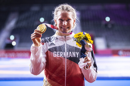 Aline Rotter-Focken, die 2020 in Tokio Gold im Ringen holte, strahlt in die Kamera und präsentiert stolz ihre Goldmedaille. Im Hintergrund sieht man sehr verschwommen das Sportstadion.