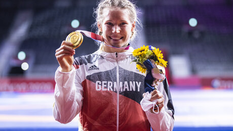 Aline Rotter-Focken, die 2020 in Tokio Gold im Ringen holte, strahlt in die Kamera und präsentiert stolz ihre Goldmedaille. Im Hintergrund sieht man sehr verschwommen das Sportstadion.