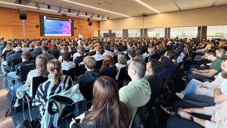 Auf dem Foto ist die bis auf den letzten Platz besetzte Aula zu sehen: Rund 1.100 Bachelor- und Masterstudierende starten gemeinsam in ihr Studium zum Wintersemester 2026/2027 und lauschen der Ansprache von Hochschulrektor Prof. Dr. Harald Riegel, der vorne am Rednerpult steht.