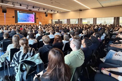 Auf dem Foto ist die bis auf den letzten Platz besetzte Aula zu sehen: Rund 1.100 Bachelor- und Masterstudierende starten gemeinsam in ihr Studium zum Wintersemester 2026/2027 und lauschen der Ansprache von Hochschulrektor Prof. Dr. Harald Riegel, der vorne am Rednerpult steht.