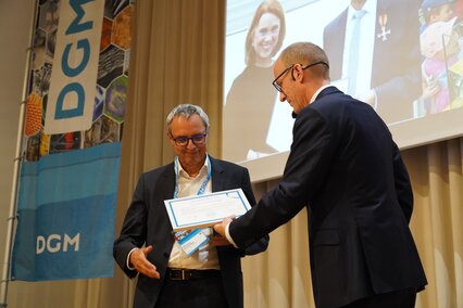 Auf dem Foto ist Prof. Dr. Gerhard Schneider zu sehen, der sehr erfreut beim DGM-Tag 2026  in Darmstadt die Ehrenmitgliedschaft der Deutschen Gesellschaft für Materialkunde aus den Händen von DGM-Präsident Dr. Till Schneiders entgegennimmt. 