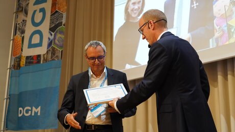 Auf dem Foto ist Prof. Dr. Gerhard Schneider zu sehen, der sehr erfreut beim DGM-Tag 2026  in Darmstadt die Ehrenmitgliedschaft der Deutschen Gesellschaft für Materialkunde aus den Händen von DGM-Präsident Dr. Till Schneiders entgegennimmt. 