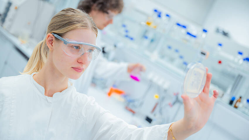 Eine Studentin prüft ein Experiment. Im Hintergrund ist ein Student zu sehen, der ein Exprimiert ausführt.