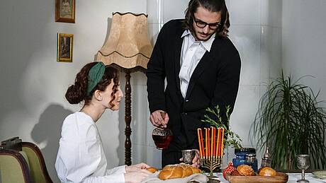 Ein junges jüdisches Paar sitzt an einem gedeckten Esstisch, auf dem ein Chanukka-Leuchter steht. dem ein 