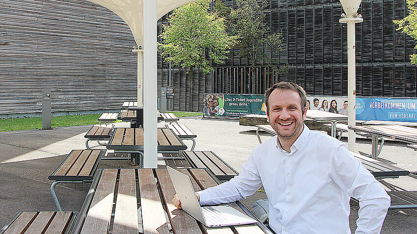Prof. Dr. Daniel Gartner sitzt mit seinem Laptop unter den Sonnenschirmen auf dem Campus Burren.