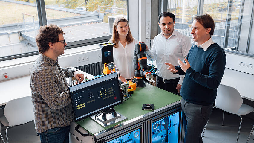 Ankuendigung Innovationsforum Auftakt KI-Challenge Vier Personen stehen um einen Roboterarm auf einem Schreibtisch.