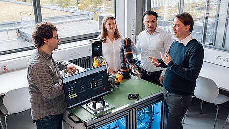 Vier Personen stehen um einen Roboterarm auf einem Schreibtisch.