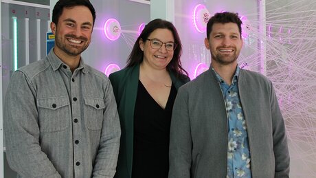 Robert Luft, Prof. Dr. Doris Aschenbrenner und Linus Hoppe stehen vor einem Exponat, das pinkfarbenen leuchtet und das veranschaulicht, wie ein neuronales Netz funktioniert. Alle drei strahlen in die Kamera und freuen sich über die Qualifizierung für die nächste Wettbewerbsrunde bei der AI-BOOST Challenge.