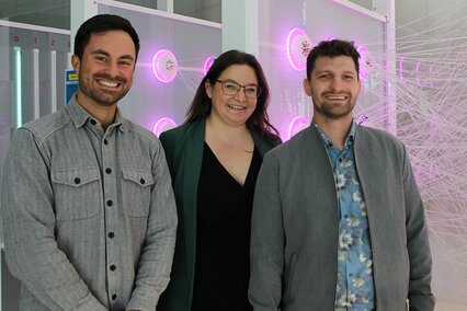 Robert Luft, Prof. Dr. Doris Aschenbrenner und Linus Hoppe stehen vor einem Exponat, das pinkfarbenen leuchtet und das veranschaulicht, wie ein neuronales Netz funktioniert. Alle drei strahlen in die Kamera und freuen sich über die Qualifizierung für die nächste Wettbewerbsrunde bei der AI-BOOST Challenge.