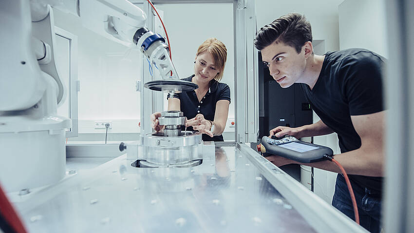 In einer Plexiglaszelle arbeiten zwei Forschende an einer Linse unterstützt von einem Industrieroboter.