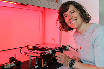 Die neue Stiftungsprofessorin für Mikrooptische Systeme, Prof. Dr. Andrea Toulouse, sitzt vor einem Versuchsaufbau und wird dabei von rötlichem Licht angestrahlt.
