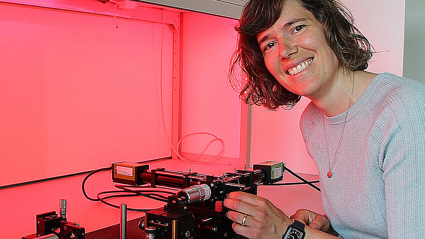Die neue Stiftungsprofessorin für Mikrooptische Systeme, Prof. Dr. Andrea Toulouse, sitzt vor einem Versuchsaufbau und wird dabei von rötlichem Licht angestrahlt.
