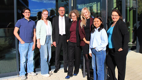 Vor dem Digital Innovation Space (DIS) stehen Adrian Rojas, Lorena Lenz (Studiengangskoordinatorin), Thilo Rentschler  (Hauptgeschäftsführer der IHK Ostwürttemberg), Prof. Dr. Anja Dieckmann (Prorektorin für Internationales), Sarah Dotschkal (MINT-Referentin Region Ostwürttemberg) Sharon Vásquez und Prof. Dr. Doris Aschenbrenner. Die Gruppe freut sich über die Auszeichnung Ostwürttembergs als „Leuchtturm-Region MINTernational“ und strahlt in die Kamera. Hinter ihr ist an der Tür die Aufschrift "Willkommen" zu sehen. 