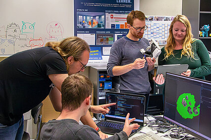 Auf dem Foto ist Prof. Dr. Marcus Gelderie mit seinem Projektteam zu sehen. Sie schauen in einen Laptop und freuen sich über das neue Lernspiel "Critical Chains".