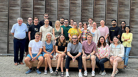 Auf dem Bild sieht man die Lehrenden und Studierenden des Masterstudiengangs Vision Science and Business (Optometry) der Hochschule Aalen zum Auftakt des Studienjahres 2026/27. Die Gruppe steht vor dem mit Holz verkleideten Studienganggebäude auf dem Campus Burren. Alle schauen fröhlich in die Kamera.