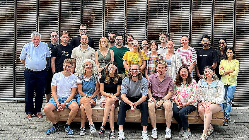 Auf dem Bild sieht man die Lehrenden und Studierenden des Masterstudiengangs Vision Science and Business (Optometry) der Hochschule Aalen zum Auftakt des Studienjahres 2026/27. Die Gruppe steht vor dem mit Holz verkleideten Studienganggebäude auf dem Campus Burren. Alle schauen fröhlich in die Kamera.