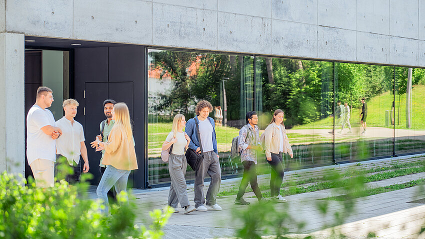 Mehrere Gruppen von Studierenden, unterhalten sich auf dem Vorplatz des Aula- / Hörsaalgebäudes.
