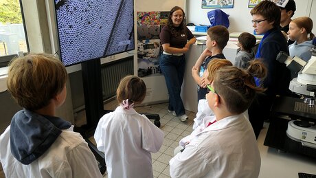 Auf dem Foto sieht man IMFAA-Doktorandin Sara Nester vor einem großen Bildschirm stehen, auf dem die mikroskopische Vergrößerung eines Insektenflügels abgebildet ist. Gebannt steht eine Gruppe von Kindern und Jugendlichen vor der jungen Frau und hört aufmerksam ihren Erklärungen über die Wunderwelt der Mikroskopie im Rahmen des Aktionstags "Türen auf mit der Maus" an der Hochschule Aalen zu.