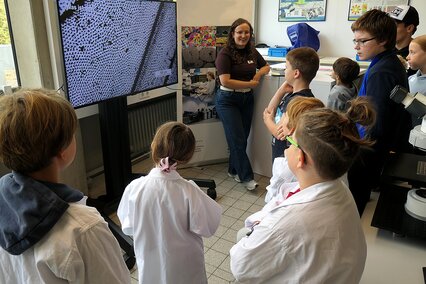 Auf dem Foto sieht man IMFAA-Doktorandin Sara Nester vor einem großen Bildschirm stehen, auf dem die mikroskopische Vergrößerung eines Insektenflügels abgebildet ist. Gebannt steht eine Gruppe von Kindern und Jugendlichen vor der jungen Frau und hört aufmerksam ihren Erklärungen über die Wunderwelt der Mikroskopie im Rahmen des Aktionstags "Türen auf mit der Maus" an der Hochschule Aalen zu.
