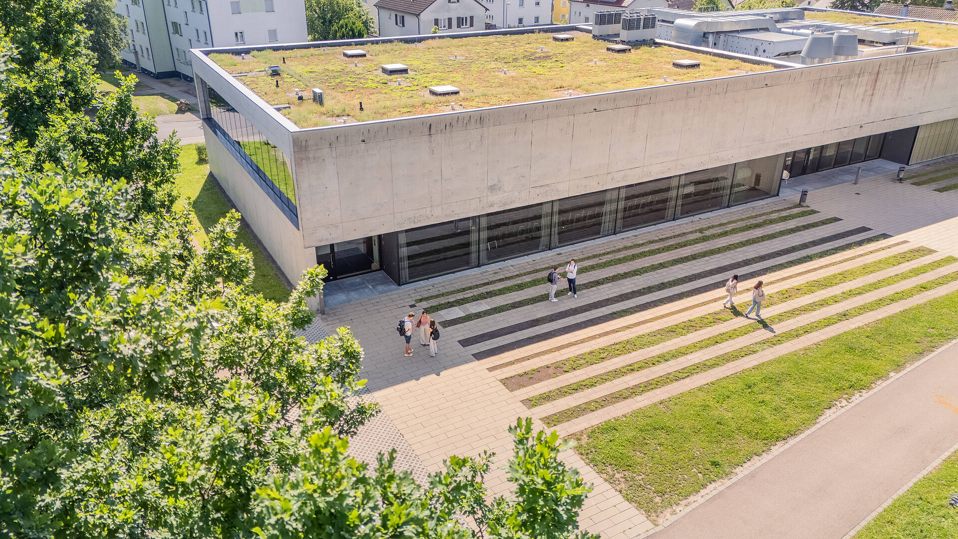 Das Aula- Hörsaalgebäude von oben. Auf dem Vorplatz unterhalten sich Studierende.