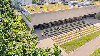 Das Aula- Hörsaalgebäude von oben. Auf dem Vorplatz unterhalten sich Studierende.
