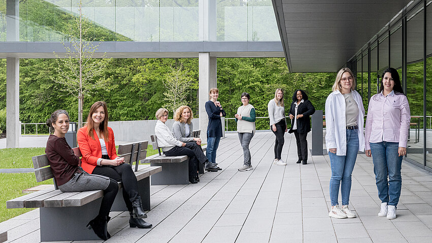 Vor einem Gebäude mit Glassteg in ein anderes Gebäude sind mehrere Frauen zu sehen, die in kleinen Grüppchen in die Kamera schauen.