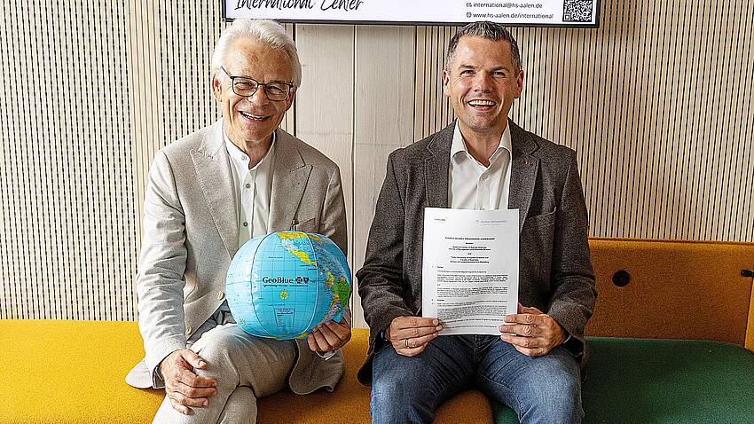 Zwei Professoren zeigen sitzend eine Urkunde und eine Weltkugel in die Kamera.