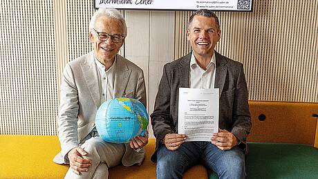 Zwei Professoren zeigen sitzend eine Urkunde und eine Weltkugel in die Kamera.