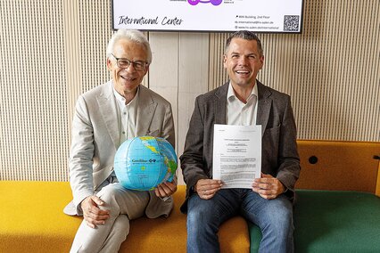 Zwei Professoren zeigen sitzend eine Urkunde und eine Weltkugel in die Kamera.