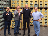 Foto: Vier grinsende Männer mit Bierflaschen vor Stapeln aus Bierkisten