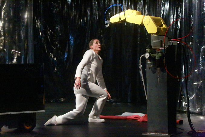 Fotografie: Eine Szene im Theater: eine Person steht ängstlich gebückt vor einem Roboterarm.