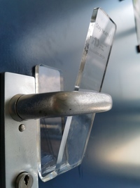  Foto: Demonstration einer Plastikapplikation an einer Klinke, die das Öffnen der Tür nur mit dem Unterarm ermöglicht.